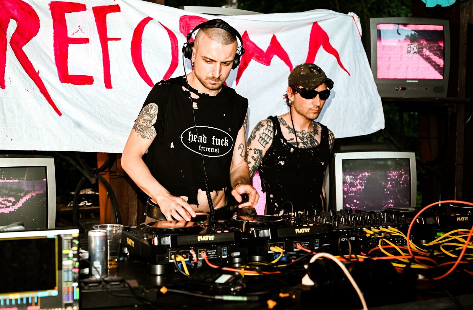 Dwie osoby DJ-skie przy konsolecie, za nimi ręcznie malowany baner i stare telewizory